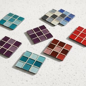 May include: A collection of square coasters, each composed of smaller, colorful tiles. The coasters feature a variety of colors, including teal, purple, red, and brown. The tiles are arranged in a grid pattern, creating a mosaic effect. The coasters are displayed on a white surface.