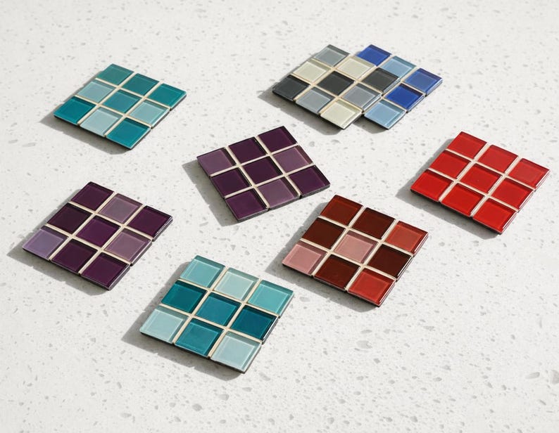 May include: A collection of square coasters, each composed of smaller, colorful tiles. The coasters feature a variety of colors, including teal, purple, red, and brown. The tiles are arranged in a grid pattern, creating a mosaic effect. The coasters are displayed on a white surface.