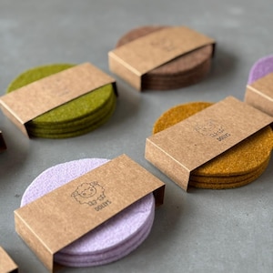 Könnte beinhalten: Eine Sammlung farbiger Filzuntersetzer in Grün-, Braun-, Lila- und Goldtönen. Jeder Stapel Untersetzer ist mit einer braunen Papphülle verpackt, die ein Schaf-Logo und das Wort "DOLLYS" trägt.