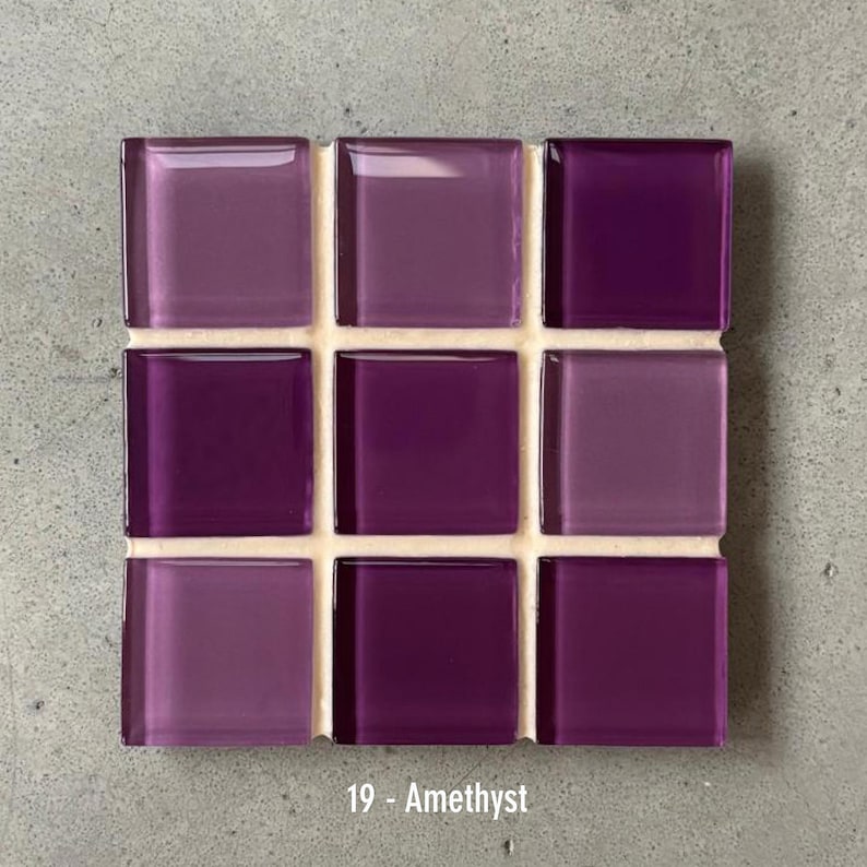 May include: A square mosaic tile sheet featuring nine glass tiles in varying shades of purple. The tiles are arranged in a 3x3 grid and are labeled "19 - Amethyst". The tiles are mounted on a white backing.