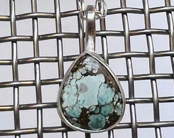 May include: A teardrop-shaped turquoise pendant with dark brown matrix veining, set in a polished silver bezel. The pendant hangs from a silver chain with oval links, showcasing a natural stone with a mottled blue and brown color palette.