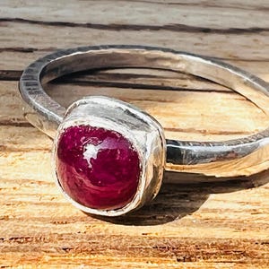 Puede incluir: Un anillo de plata con una gema grande, ovalada y de color rojo intenso. La banda del anillo tiene un diseño texturizado, y la gema está engastada en un bisel liso y pulido. El anillo se exhibe sobre una superficie de madera.