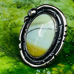 Könnte beinhalten: Ovaler Ring mit silberfarbener Fassung. Der Ring hat einen hellgrünen und gelben Stein mit dunklen Linien. Die Fassung hat ein dekoratives, strukturiertes Design. Der Ring ist vor einem grünen Hintergrund platziert.