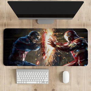 Puede incluir: Alfombrilla de escritorio rectangular con una ilustración dinámica de Capitán América e Iron Man en batalla. La imagen muestra a los héroes enfrentándose, con chispas y efectos de energía. Se ven un teclado, un ratón y un monitor.