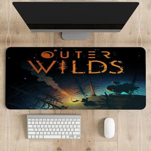 Pode incluir: Tapete de rato retangular preto com o texto laranja "OUTER WILDS" e uma ilustração de uma cena de floresta e uma fogueira. Um monitor de computador, um teclado e um rato são visíveis acima e abaixo.