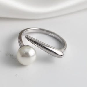 May include: A silver ring featuring a large, round, white pearl and a streamlined, curved design. The ring has a modern, minimalist style. The band is marked with "S925". A stylish piece of jewellery.
