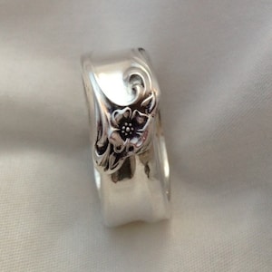 May include: A silver ring with a floral design. The ring features a detailed flower and leaf pattern on the side. The ring is made of a shiny, reflective metal and is set against a soft, white background.