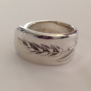 May include: A silver ring with a wheat design. The ring is wide and has a rounded shape.