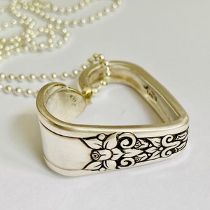 May include: A silver heart-shaped pendant necklace made from a repurposed spoon. The spoon has a floral design and is attached to a silver ball chain.