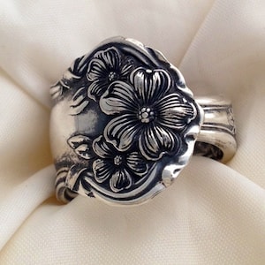 May include: A silver spoon ring with a floral design. The ring features three flowers with five petals each, and the flowers are surrounded by leaves. The ring is made from a vintage spoon.