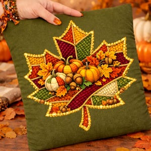 Op de afbeelding: Groen vierkant kussen met een gedetailleerd geborduurd ontwerp van een herfstblad. Het blad is gevuld met pompoenen, eikels en herfstbladeren in oranje, gele en rode tinten. Het kussen heeft een gouden rand.