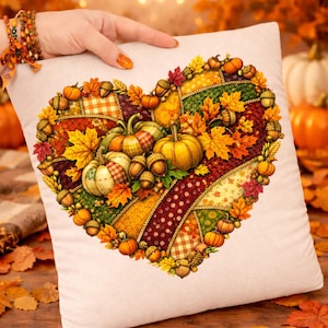 Op de afbeelding: Een vierkant, crèmekleurig kussen met een hartvormig ontwerp. Het hart bestaat uit patchworkpatronen, pompoenen, eikels en herfstbladeren in oranje, gele en rode tinten. Het kussen wordt door een persoon vastgehouden.