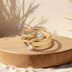 Può includere: Un anello color oro con un design a spirale, con due pietre preziose rotonde e trasparenti. L'anello è esposto su un piedistallo di legno con uno sfondo chiaro, creando un'estetica minimalista. Il design dell'anello è moderno ed elegante.