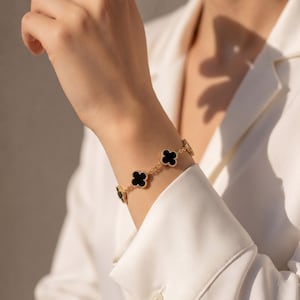 Op de afbeelding: Een gouden kettingarmband met meerdere zwarte klavervormige bedels. De armband bestaat uit een fijne gouden schakelketting die verschillende symmetrische vierbladig klavermotieven verbindt, elk ingelegd met een glad, zwart materiaal.