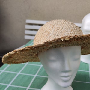 Può includere: Un cappello di paglia naturale con tesa larga, esposto su una testa di manichino bianca. Il cappello è di colore marrone chiaro e ha un bordo leggermente sfilacciato. Il cappello è su una superficie a scacchi verdi e bianchi.