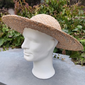 May include: A woven straw hat with a wide brim, displayed on a white mannequin head. The hat is a natural beige color, with a slightly darker rim. The hat's brim is approximately 15 inches in diameter, offering sun protection. The hat is suitable for outdoor wear.