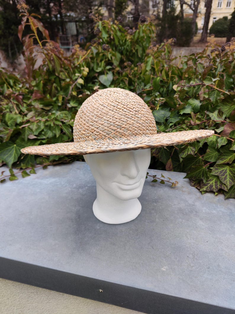 May include: A woven straw hat with a wide brim and a rounded crown. The hat is displayed on a white mannequin head against a backdrop of green foliage. The hat's natural color and texture are prominent.