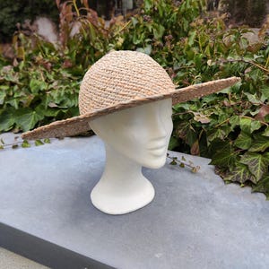 May include: A natural straw hat with a wide brim, displayed on a white mannequin head. The hat has a woven texture and a rounded crown. The brim extends outwards, providing shade. The background features green foliage.