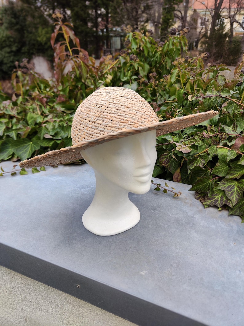 May include: A natural straw hat with a wide brim, displayed on a white mannequin head. The hat has a woven texture and a rounded crown. The brim extends outwards, providing shade. The background features green foliage.