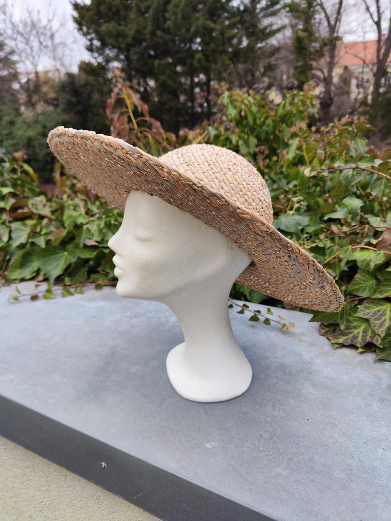 May include: A wide-brimmed straw hat with a natural woven texture. The hat is displayed on a white mannequin head, showcasing its shape and design. The hat's brim is wide, providing ample shade. The hat is a fashion accessory.