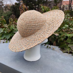 May include: A woven straw hat with a wide brim and a rounded crown. The hat is displayed on a white mannequin head. The hat's natural beige color complements the outdoor setting, suggesting a summer accessory.