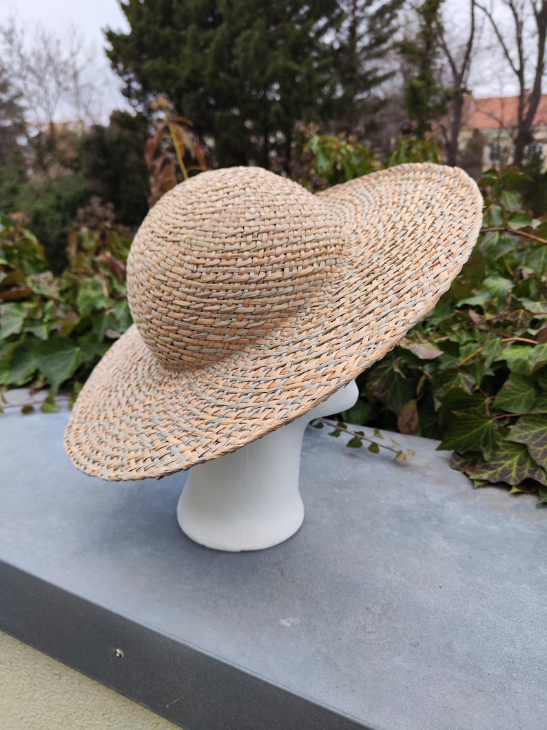 May include: A woven straw hat with a wide brim and a rounded crown. The hat is displayed on a white mannequin head. The hat's natural beige color complements the outdoor setting, suggesting a summer accessory.