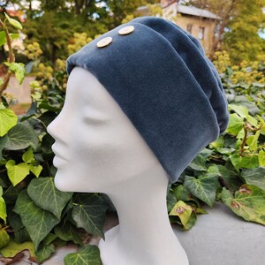 May include: A teal velvet beanie hat with two gold-colored buttons. The hat is displayed on a white mannequin head against a backdrop of green foliage and a blurred outdoor scene.