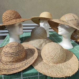 May include: A collection of woven straw hats in various styles, including wide-brimmed sun hats and smaller, more structured designs. The hats are displayed on white mannequin heads, showcasing their shape and texture. The hats are a natural beige color.