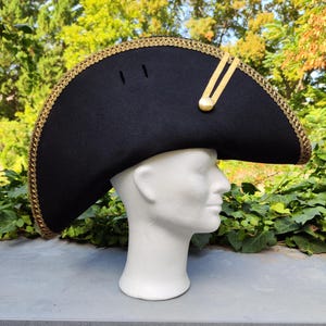 Op de afbeelding: Een zwarte tricornhoed met een gouden galonversiering en een gouden knoop. De hoed is schuin naar de zijkant gekanteld en heeft een lichte kromming aan de rand.