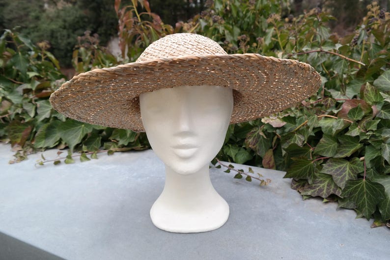 May include: A wide-brimmed, woven straw hat with a rounded crown. The hat is a natural beige color and sits atop a white mannequin head. The background features green foliage and a light gray surface.