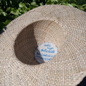 May include: A wide-brimmed straw hat with a woven texture. The hat features a circular label with the word "wellenkind" and contact information in blue text. The hat is set against a backdrop of green foliage.