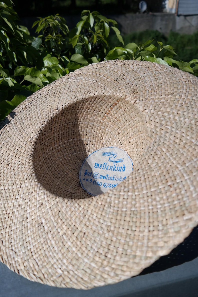 May include: A wide-brimmed straw hat with a woven texture. The hat features a circular label with the word "wellenkind" and contact information in blue text. The hat is set against a backdrop of green foliage.