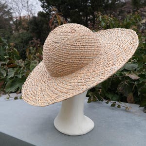 May include: A woven straw hat with a wide brim and a rounded crown. The hat is displayed on a white mannequin head. The hat's natural beige color contrasts with the green foliage in the background. This hat is a stylish accessory.