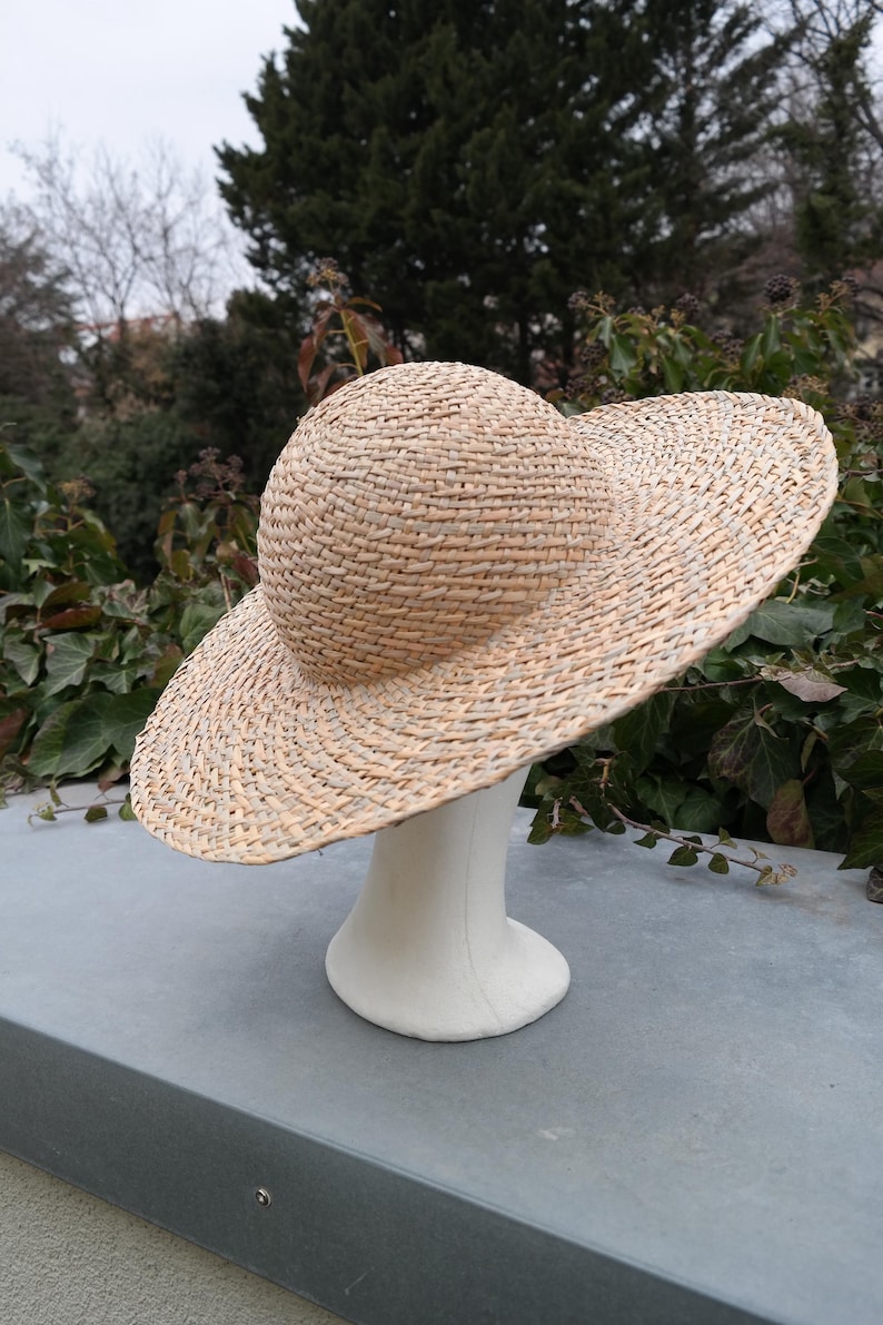 May include: A woven straw hat with a wide brim and a rounded crown. The hat is displayed on a white mannequin head. The hat's natural beige color contrasts with the green foliage in the background. This hat is a stylish accessory.
