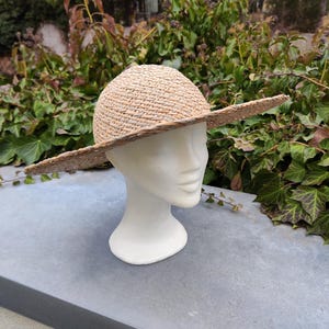 May include: A natural straw hat with a wide brim, displayed on a white mannequin head. The hat has a rounded crown and a broad, flat brim, perfect for sun protection. The hat is made of woven straw.