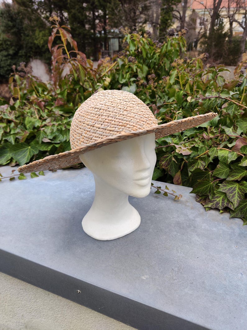 May include: A natural straw hat with a wide brim, displayed on a white mannequin head. The hat has a rounded crown and a broad, flat brim, perfect for sun protection. The hat is made of woven straw.