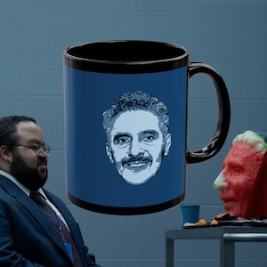 May include: A black coffee mug with a blue background and a white illustration of a man's face with a mustache. The mug has a black handle.