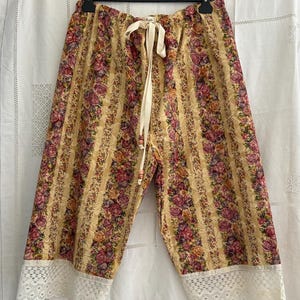 Può includere: Un paio di pantaloni a metà polpaccio con un motivo a righe verticali che alterna fasce di stampe floreali nei toni del rosa, viola e giallo su sfondo beige. I pantaloni presentano una coulisse color crema in vita e un bordo in pizzo bianco.