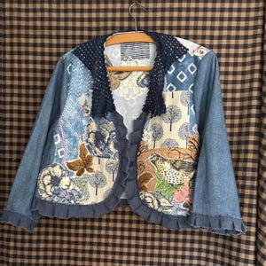 May include: A patchwork denim jacket with a unique design. The jacket features various fabric patches with floral, butterfly, and bird motifs. It has a navy blue crochet collar and ruffled edges, creating a bohemian aesthetic. The jacket is hanging on a wooden hanger.