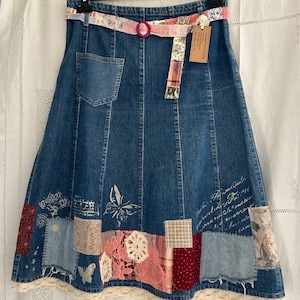 May include: A blue denim skirt featuring a patchwork hem with various fabric squares, lace accents, and butterfly motifs. The skirt includes a matching fabric belt with a pink ring buckle, a single back pocket, and a cream-coloured lace trim at the bottom.