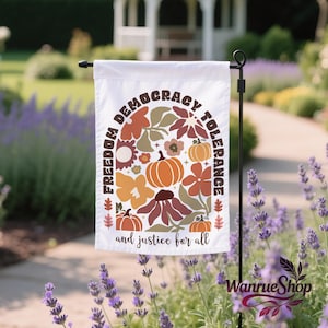 Könnte beinhalten: Weiße Gartenflagge mit den Worten "FREEDOM DEMOCRACY TOLERANCE" in einem Halbkreis, darunter "and justice for all". Die Flagge zeigt ein Blumendesign mit Kürbissen und Blumen in Orange, Braun und Gelb.