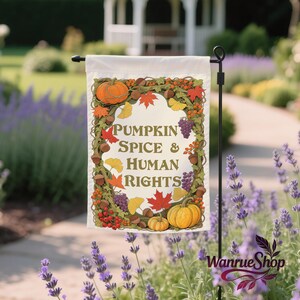Könnte beinhalten: Eine weiße Gartenflagge mit dem Text "PUMPKIN SPICE & HUMAN RIGHTS", umgeben von einem Kranz aus Herbstlaub, Kürbissen und Trauben. Die Flagge wird auf einem schwarzen Metallständer in einem Garten präsentiert.