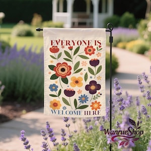May include: A cream garden flag with the text "EVERYONE IS WELCOME HERE" in red and blue. The flag features a floral design with various colourful flowers and green leaves. The flag is displayed on a black metal stand in a garden setting.