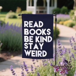 May include: A navy blue garden flag with the words "READ BOOKS BE KIND STAY WEIRD" in white block letters. The flag is displayed outdoors with a black metal stand, surrounded by lavender plants and a white gazebo in the background.