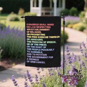 May include: Black garden flag with the First Amendment text in rainbow colors. The flag is displayed outdoors in a garden setting with lavender plants and a white gazebo in the background. The flag reads: Congress shall make no law respecting an establishment of religion.