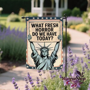 May include: A garden flag featuring the Statue of Liberty holding a sign that reads "WHAT FRESH HORROR DO WE HAVE TODAY?" The flag has a vintage design with a blue and red border and star accents. The flag is displayed outdoors.