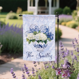 May include: A light blue garden flag featuring a floral design with blue and white hydrangeas in a decorative vase. The flag has a damask pattern background and the text "The Smiths" in a script font.