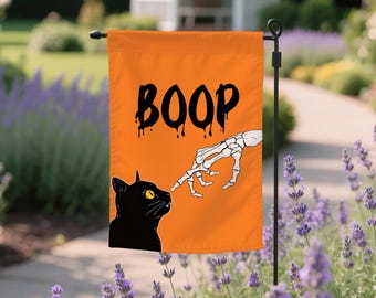 May include: Orange Halloween garden flag with the word "BOOP" in black dripping font. A black cat with yellow eyes is being touched by a skeleton hand. The flag is displayed outdoors.
