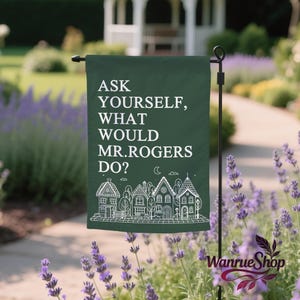 May include: A dark green garden flag featuring the text "ASK YOURSELF, WHAT WOULD MR. ROGERS DO?" in white. A line drawing of houses and trees is at the bottom. The flag is displayed outdoors in a garden setting.