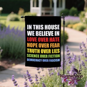 May include: Black garden flag with white text: "IN THIS HOUSE WE BELIEVE IN LOVE OVER HATE HOPE OVER FEAR TRUTH OVER LIES SCIENCE OVER FICTION DEMOCRACY OVER FASCISM." The flag is displayed outdoors, surrounded by lavender.